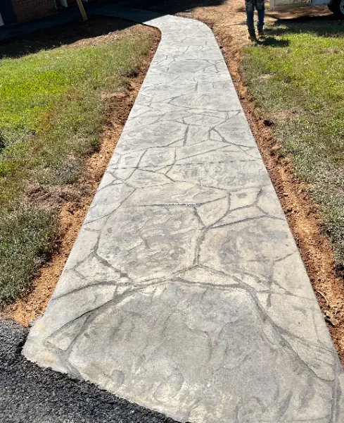 Stamped concrete walkway curving from an asphalt edge toward a house, with grass and bare soil along both sides.