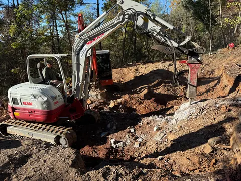 Excavator running a hydraulic breaker through rock on a hillside cut.